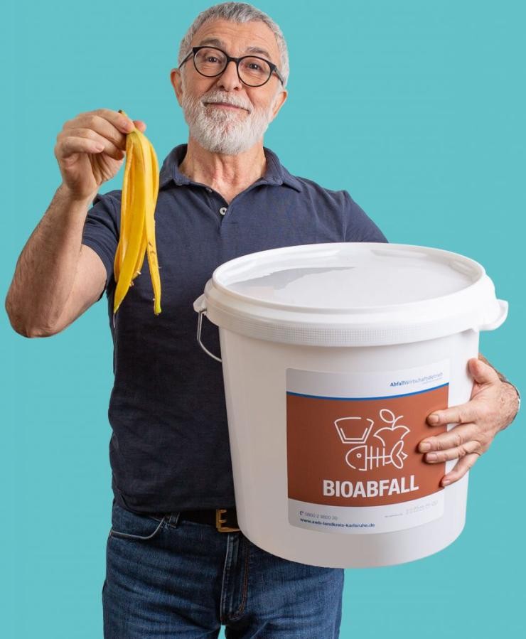 Das Bringsystem Ein Mann hält mit der rechten Hand eine Bananenschale hoch mit dem linken Arm umfasst er eine weißen Eimer für Bioabfall.