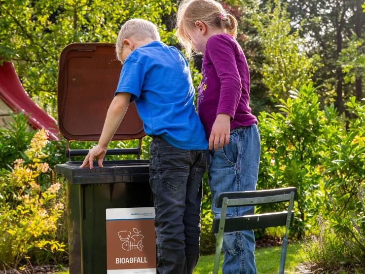 Eine graue Biotonne mit braunem Deckel steht im Garten. Zwei Kinder, ein Junge und ein Mädchen, schauen interessiert hinein. Zwei Kinder schauen in eine Biotonne, die im Garten steht.