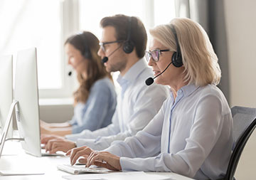 Drei Personen mit Headset sitzen vor Computern am Schreibtisch.