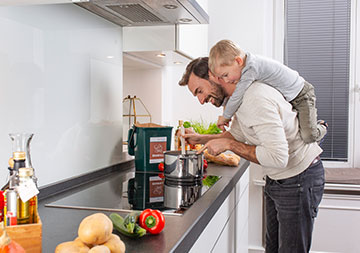 Ein Mann in der Küche und kocht. Auf seinem Rücken trägt er einen kleinen Jungen.