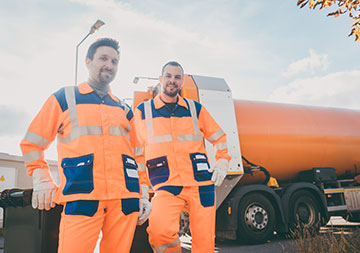Zwei Müllwerker stehen vor einem Müllfahrzeug; im Hintergrund blauer Himmel.